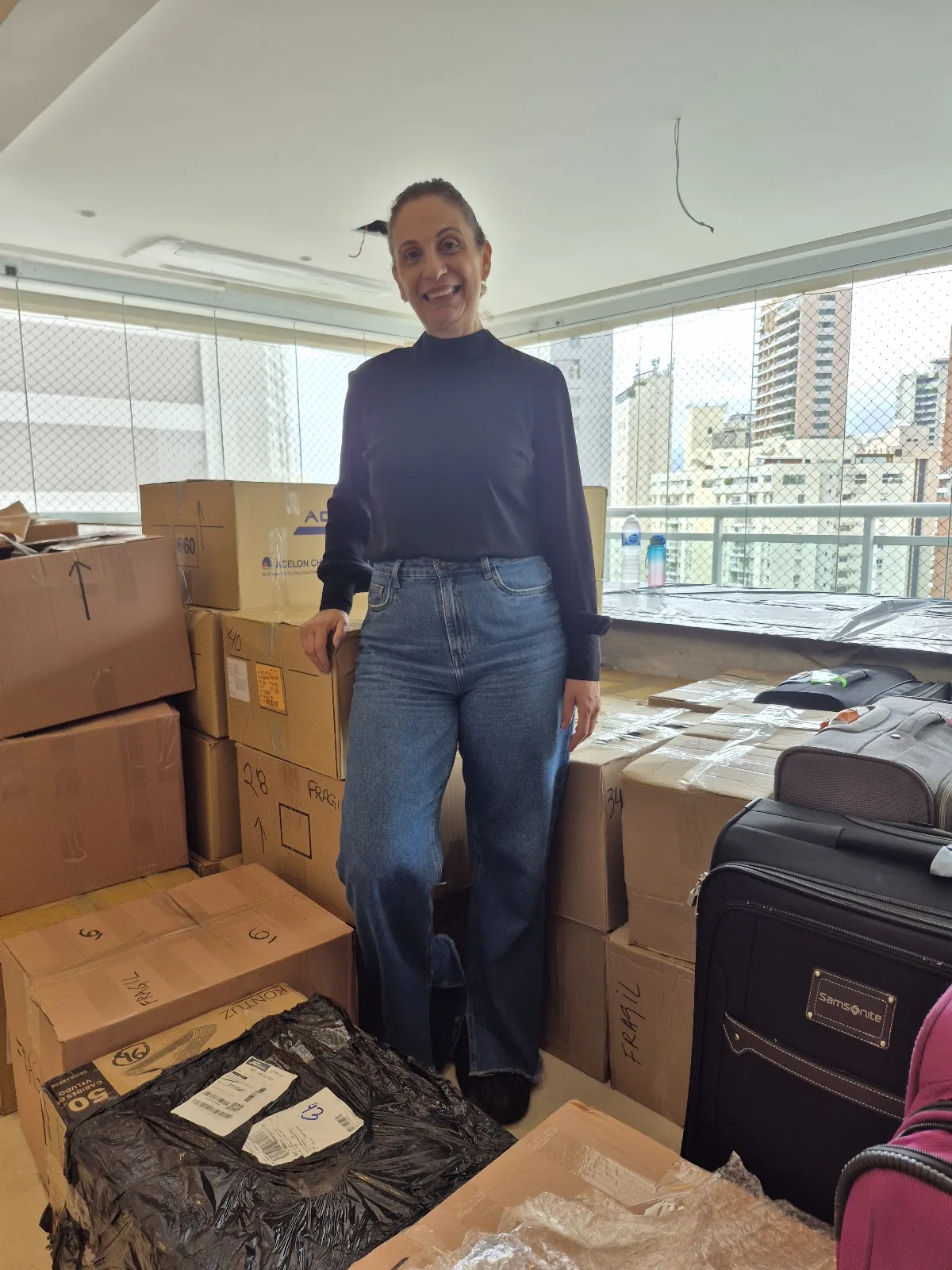 Katia sorrindo entre as caixas de uma mudança, em um apartamento com vista para a cidade